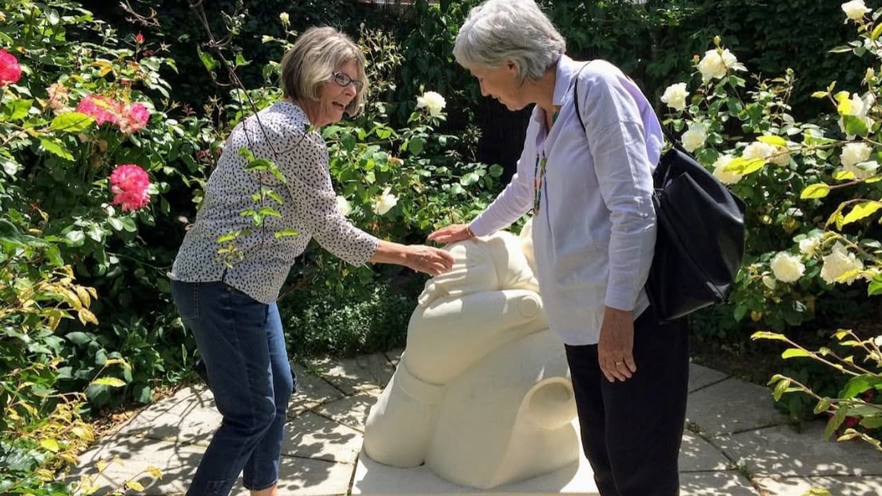 Two members admiring a carved stone sculpture in a sunlit rose garden